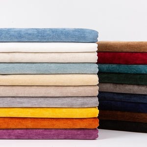 Op de afbeelding: Stapels kleurrijke stofstalen in diverse tinten, van lichtblauw tot dieppaars. De stoffen zijn netjes opgestapeld en tonen een verscheidenheid aan texturen en kleuren, ideaal voor bekleding of knutselprojecten.