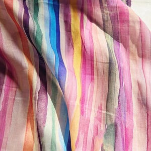 Op de afbeelding: Een close-up van een stof met verticale strepen in aquarel-stijl. De strepen hebben een levendig kleurenspectrum, waaronder roze, paars, blauw, groen, geel en oranje. De stof lijkt zacht en vloeiend, met een enigszins doorschijnende kwaliteit.