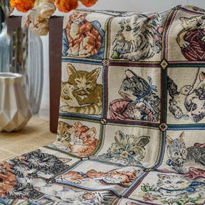 Puede incluir: Un tapiz beige con un patrón repetido de ilustraciones de gatos en marcos cuadrados. Los gatos se representan en diferentes poses y colores, con detalles como lazos y cestas. El tapiz está drapeado sobre una superficie.