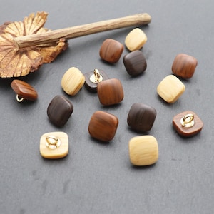 May include: A collection of square wooden buttons in various shades of brown and tan, with gold-colored metal loops for sewing. The buttons are arranged on a dark gray surface, showcasing their natural wood grain.