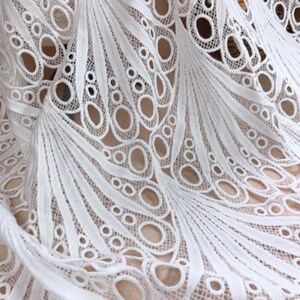 May include: Close-up of white lace fabric with a detailed, openwork design. The fabric features a repeating pattern of curved lines and circular cutouts, creating a delicate and airy texture. The lace is likely used for apparel or home decor.