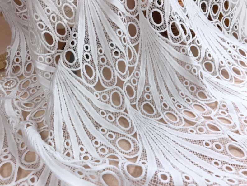 May include: Close-up of white lace fabric with a detailed, openwork design. The fabric features a repeating pattern of curved lines and circular cutouts, creating a delicate and airy texture. The lace is likely used for apparel or home decor.