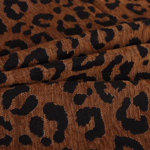 Puede incluir: Una tela texturizada con un patrón de leopardo repetitivo en tonos marrón y negro. El material tiene un tejido acanalado visible y está doblado para crear sombras suaves y profundas, además de reflejos en la superficie del textil.