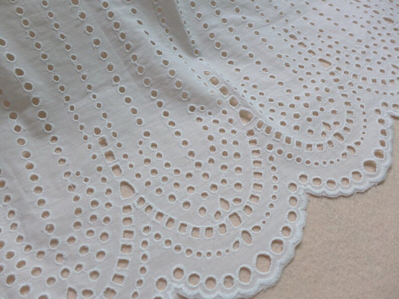 May include: White cotton lace trim with a scalloped edge and a delicate eyelet pattern. The lace has a repeating design of small circles and ovals.