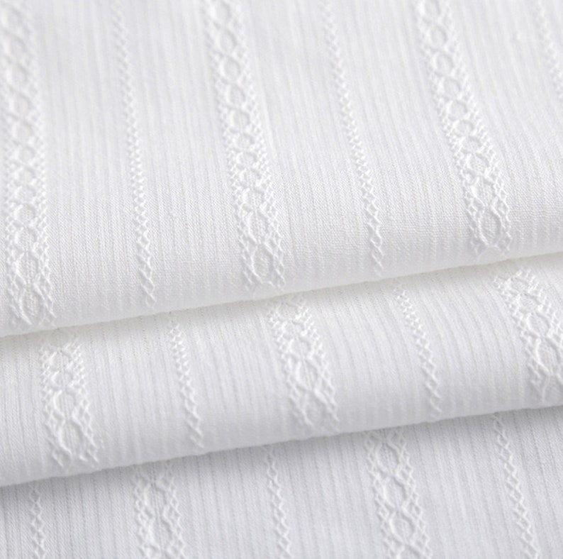 May include: White fabric with a textured, woven pattern. The fabric has vertical stripes and a delicate, raised design.