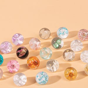 May include: A collection of spherical resin beads, each containing embedded dried flowers, petals, or small colorful stone fragments. The beads are clear and vary in color, featuring shades of pink, blue, orange, white, and black.
