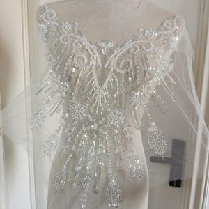 May include: A white tulle fabric appliqué featuring intricate embroidery, silver sequins, and white seed beads arranged in a symmetrical, ornate pattern with swirling lines and dangling beaded accents that extend downwards from the neckline area.