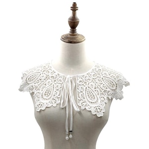 Könnte beinhalten: Ein weißer Spitzenkragen mit einem detaillierten Blumenmuster und einem Bindeband. Der Kragen, der auf einer Schaufensterpuppe präsentiert wird, hat einen gewellten Rand und zwei kleine Perlen an den Enden der Bänder. Ein Accessoire im Vintage-Stil.