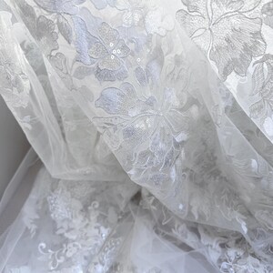 May include: White sheer fabric with a delicate floral pattern embroidered with sequins. The fabric is soft and flowing, making it ideal for a variety of projects.