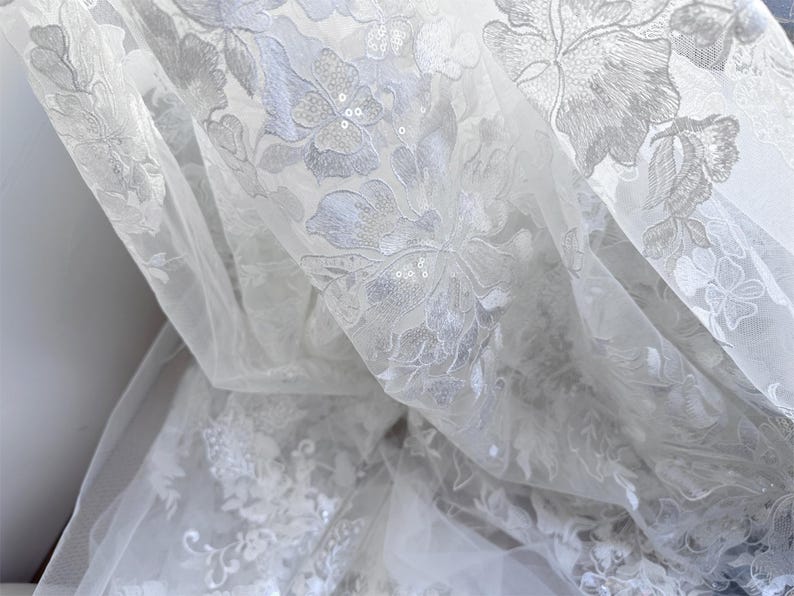 May include: White sheer fabric with a delicate floral pattern embroidered with sequins. The fabric is soft and flowing, making it ideal for a variety of projects.