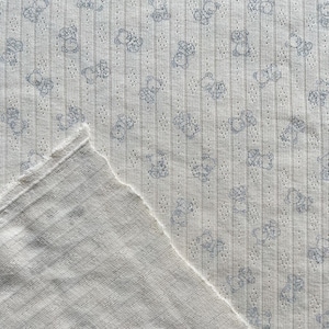 May include: Close-up of a white fabric with a repeating pattern of light blue teddy bears. The fabric has a ribbed texture with small holes and a raw edge, suggesting it may be a cut piece of material.
