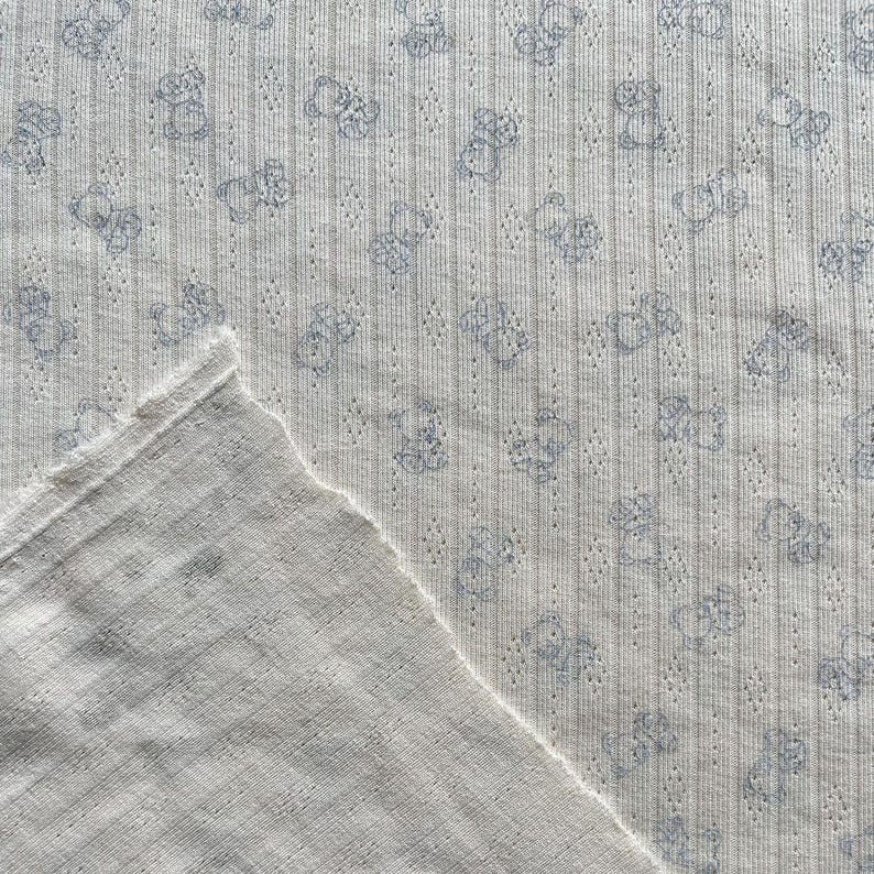 May include: Close-up of a white fabric with a repeating pattern of light blue teddy bears. The fabric has a ribbed texture with small holes and a raw edge, suggesting it may be a cut piece of material.