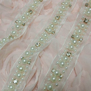 May include: White pearl and rhinestone trim ribbon. The ribbon is on a pink fabric background.