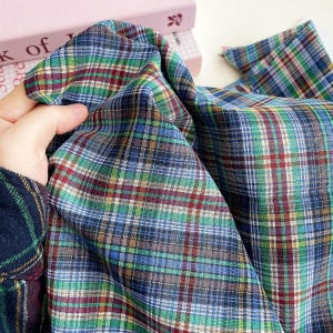 May include: A piece of fabric featuring a multi-colored plaid pattern with intersecting lines in shades of blue, green, red, and white. The textile has a visible woven texture and is draped to show the depth and complexity of the tartan design.