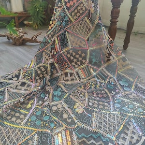 May include: Black fabric with a patchwork design featuring geometric shapes, stars, and floral patterns. The fabric is embellished with colorful sequins and embroidery in shades of pink, yellow, blue, and gold. The fabric is draped over a surface.