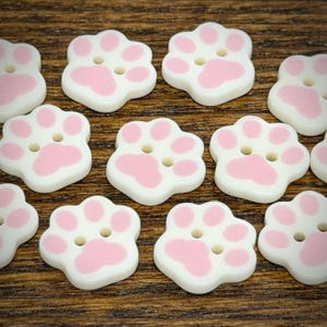 May include: A collection of white buttons shaped like animal paw prints, each featuring four holes for attachment and light pink accents on the toe pads and the main pad. The buttons have a smooth, glossy finish and are arranged in a scattered pattern.