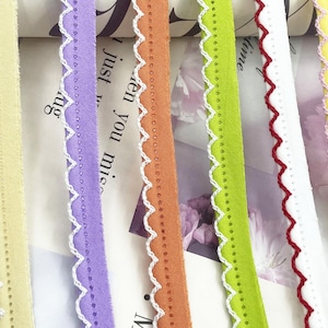 May include: Close-up of colorful ribbons with scalloped edges. The ribbons are in shades of yellow, purple, orange, green, white, and pink. Each ribbon has a white decorative trim along the edge. The ribbons are arranged vertically.