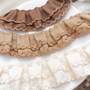Puede incluir: Primer plano de tres tiras de tela con volantes en diferentes tonos de marrón y crema. Cada tira presenta un borde festoneado y una costura decorativa en la parte inferior. La tela parece suave y ligera, adecuada para proyectos de costura.