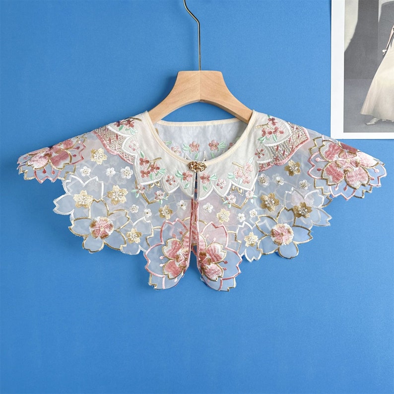 May include: A white lace collar with pink and gold floral embroidery. The collar is on a hanger against a blue background.