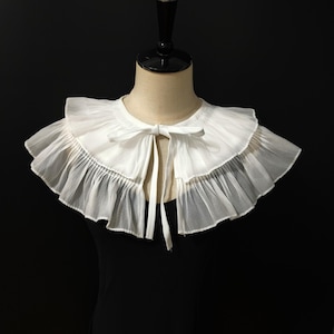 May include: A white detachable collar made of sheer, lightweight fabric featuring two layers of ruffled trim. The collar has a round neckline and is secured at the front with a matching white ribbon tie that hangs down in two long, thin strips.