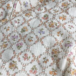 May include: Cream-colored fabric with a repeating floral pattern. The design features small, colorful flowers in shades of pink, yellow, and purple, arranged within a lattice of interconnected circles. The fabric has a slightly textured appearance.
