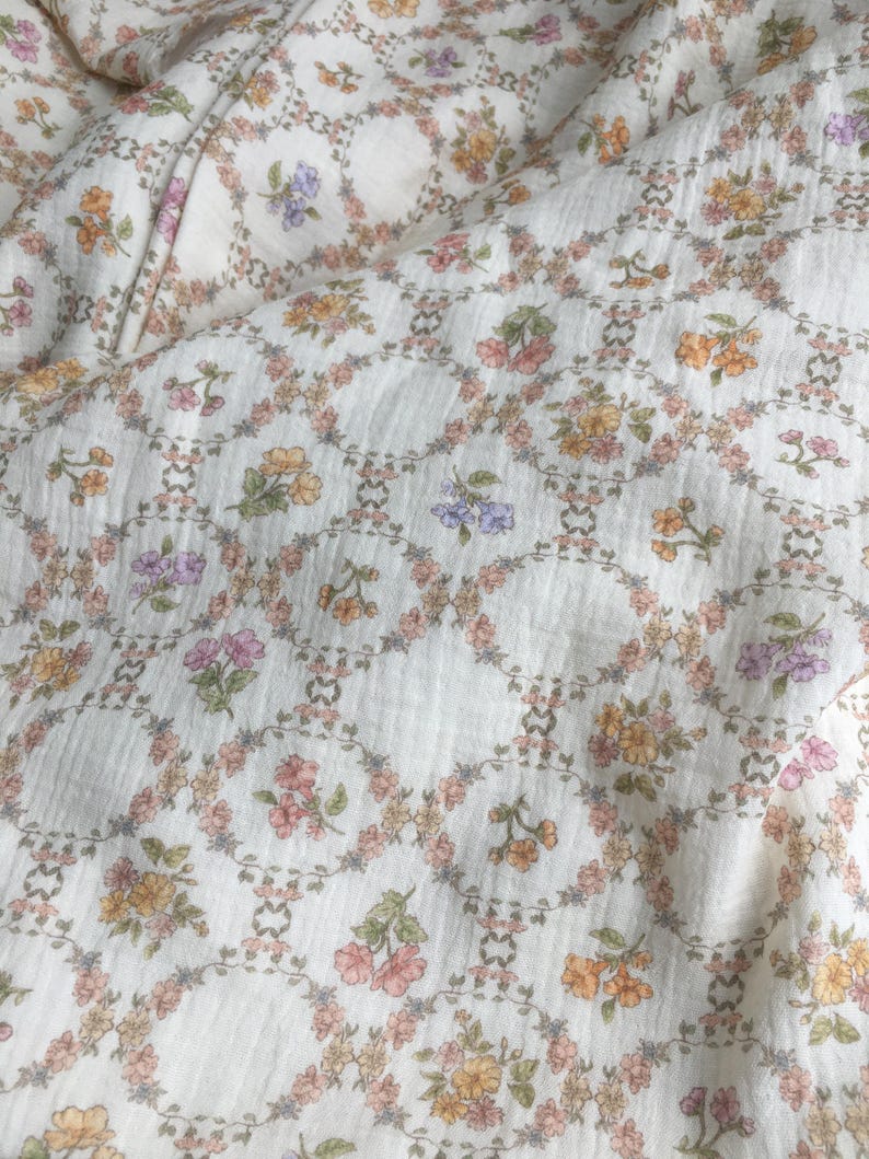 May include: Cream-colored fabric with a repeating floral pattern. The design features small, colorful flowers in shades of pink, yellow, and purple, arranged within a lattice of interconnected circles. The fabric has a slightly textured appearance.