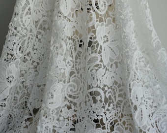 May include: Close-up of white lace fabric with a floral pattern. The delicate lace features intricate designs of flowers and leaves, creating a textured and elegant appearance. The fabric is likely used for apparel or home decor.