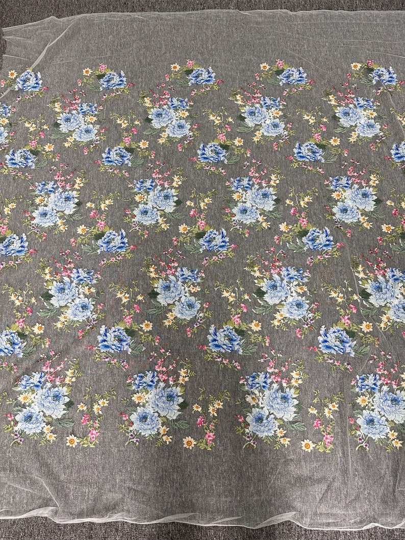Puede incluir: Tela de tul blanco con estampado floral con flores azules, rosas y amarillas. La tela tiene una textura delicada y transparente.