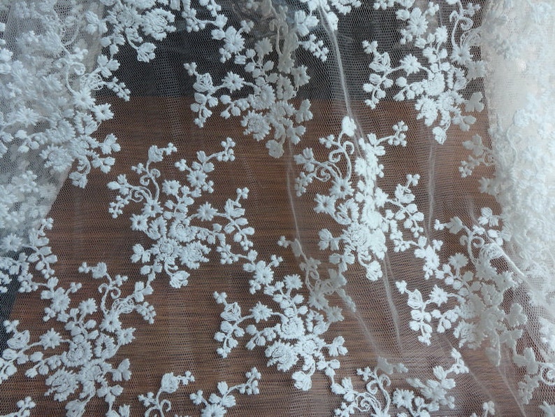 Op de afbeelding: Wit kant met een delicaat bloemenpatroon. De kant is doorschijnend en heeft een zachte, vloeiende textuur.