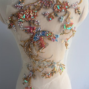 May include: A sheer nude mesh fabric applique featuring intricate embroidery and beadwork. The design depicts a bird with iridescent pink, blue, and gold sequins, metallic silver threads, and small white beads arranged in a floral and avian pattern.