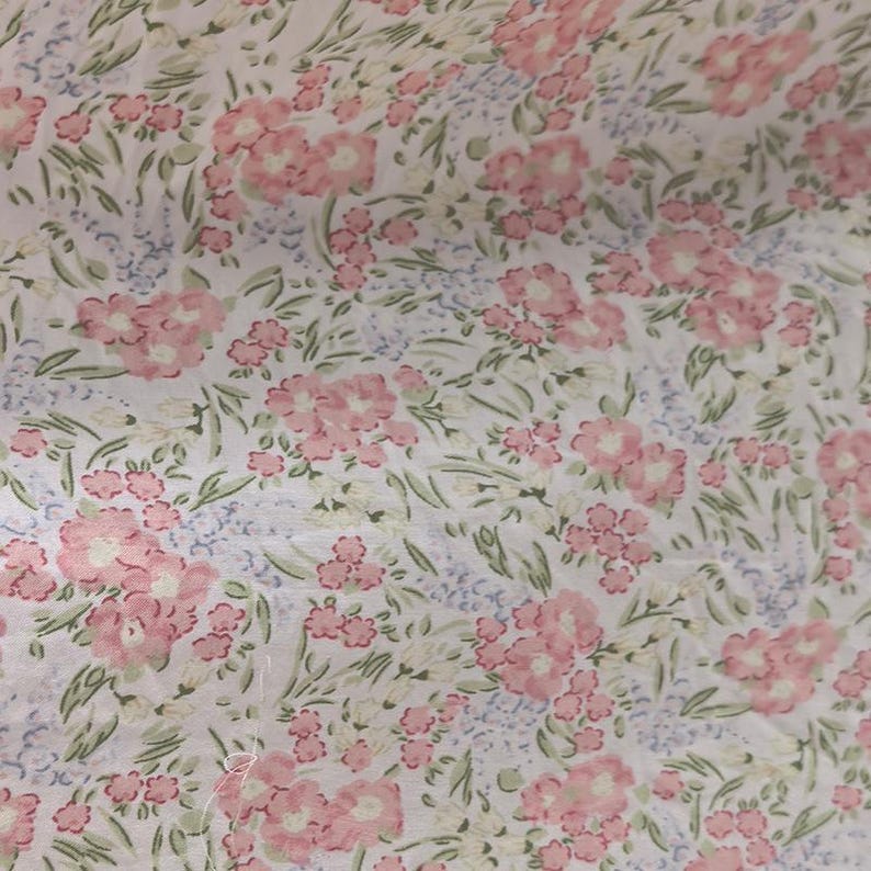 Puede incluir: Tela floral con un patr&oacute;n repetido de flores rosas, peque&ntilde;as flores azules y hojas verdes sobre un fondo blanco. La tela tiene una est&eacute;tica delicada y vintage, adecuada para diversos proyectos de costura y artesan&iacute;a.