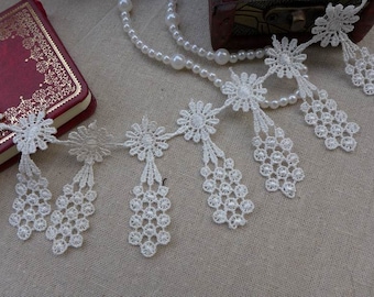 Peut inclure: Garniture en dentelle blanche avec un bord festonné et des motifs floraux délicats. La garniture présente une série de glands pendants avec des motifs complexes.