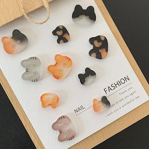 May include: A collection of ten cat-shaped buttons in various colors including orange, grey, black, and white, arranged on a white display card. The card features the printed text FASHION, NAIL, Thank you, hope to see you again, and i wish you a wonderful day.