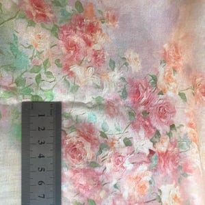 May include: Close-up of fabric with a floral pattern featuring pink, peach, and white roses with green leaves. A metal ruler is placed on the fabric, showing measurements in inches. The fabric has a soft, watercolor-like appearance.