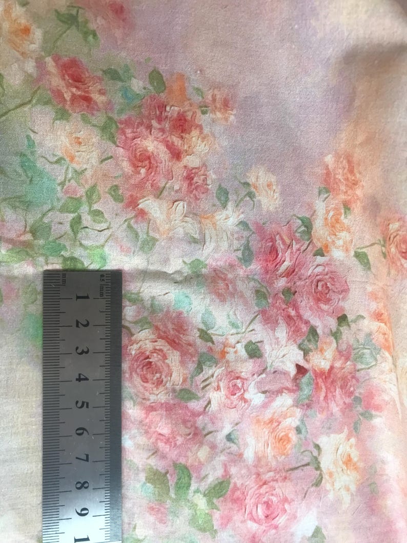 May include: Close-up of fabric with a floral pattern featuring pink, peach, and white roses with green leaves. A metal ruler is placed on the fabric, showing measurements in inches. The fabric has a soft, watercolor-like appearance.