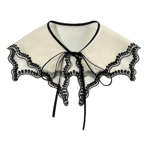 Könnte beinhalten: Ein cremefarbener, abnehmbarer Kragen mit schwarzem Besatz und einem zarten, gewellten Spitzenrand. Der Kragen wird vorne mit schwarzen Bändern gebunden. Ein stilvolles Accessoire, um jedem Outfit einen Hauch von Eleganz zu verleihen.