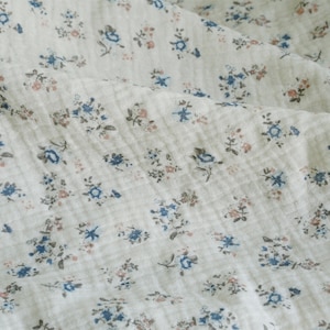 May include: A close-up of a white fabric with a small blue and pink floral pattern. The fabric has a textured, slightly wrinkled appearance.