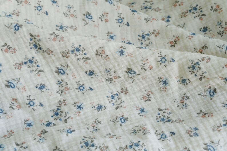 May include: A close-up of a white fabric with a small blue and pink floral pattern. The fabric has a textured, slightly wrinkled appearance.