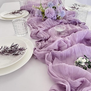 Puede incluir: Una mesa decorada con un camino de mesa de tela color lavanda con volantes. Platos blancos, vasos transparentes y pequeños arreglos florales están dispuestos sobre el mantel blanco. Acentos florales secos y un pequeño candelabro completan el look.