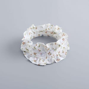 May include: A white, ruffled collar with a floral pattern in shades of pink, blue, and green. The collar has a button closure at the front and is designed to be worn around the neck. The fabric appears to be lightweight.