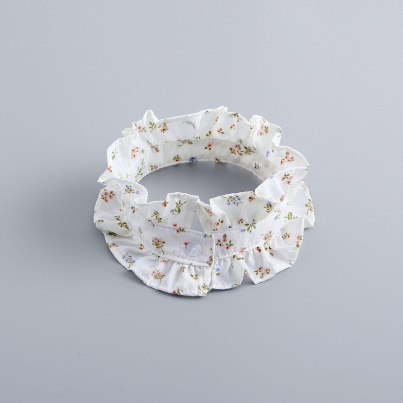May include: A white, ruffled collar with a floral pattern in shades of pink, blue, and green. The collar has a button closure at the front and is designed to be worn around the neck. The fabric appears to be lightweight.