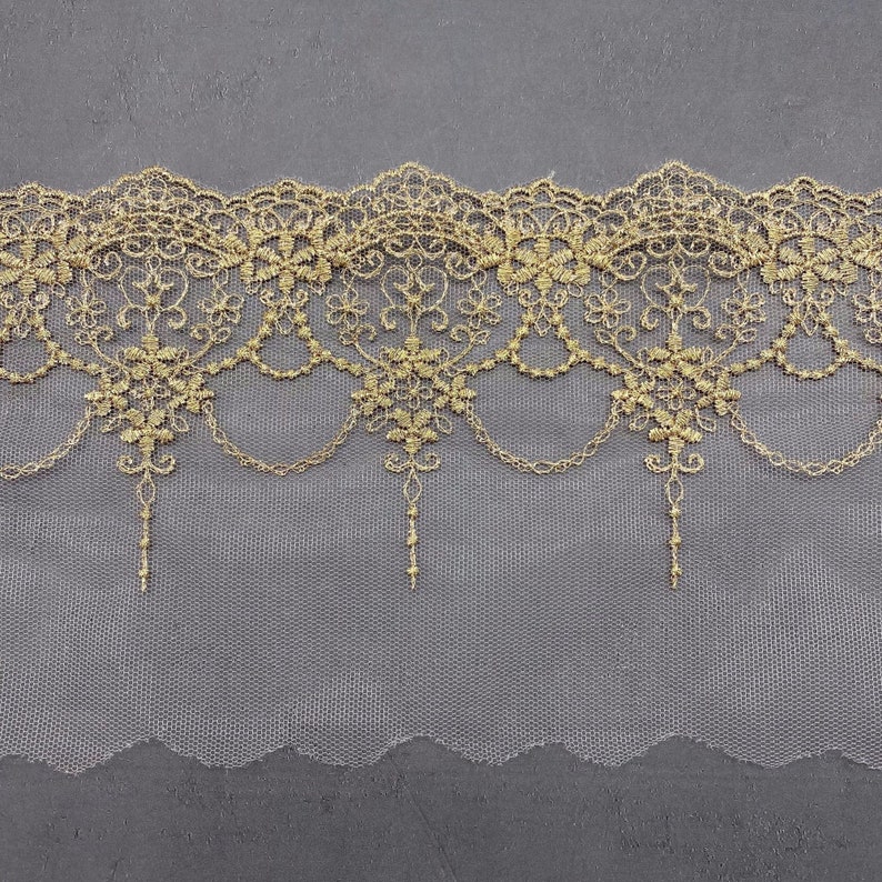 May include: Gold lace trim with a delicate floral pattern and a scalloped edge. The lace is on a sheer white mesh background.