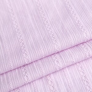 May include: Close-up of a soft, pale pink fabric with a ribbed texture and a delicate, woven pattern. The fabric is folded in layers, showcasing the intricate details of the design.