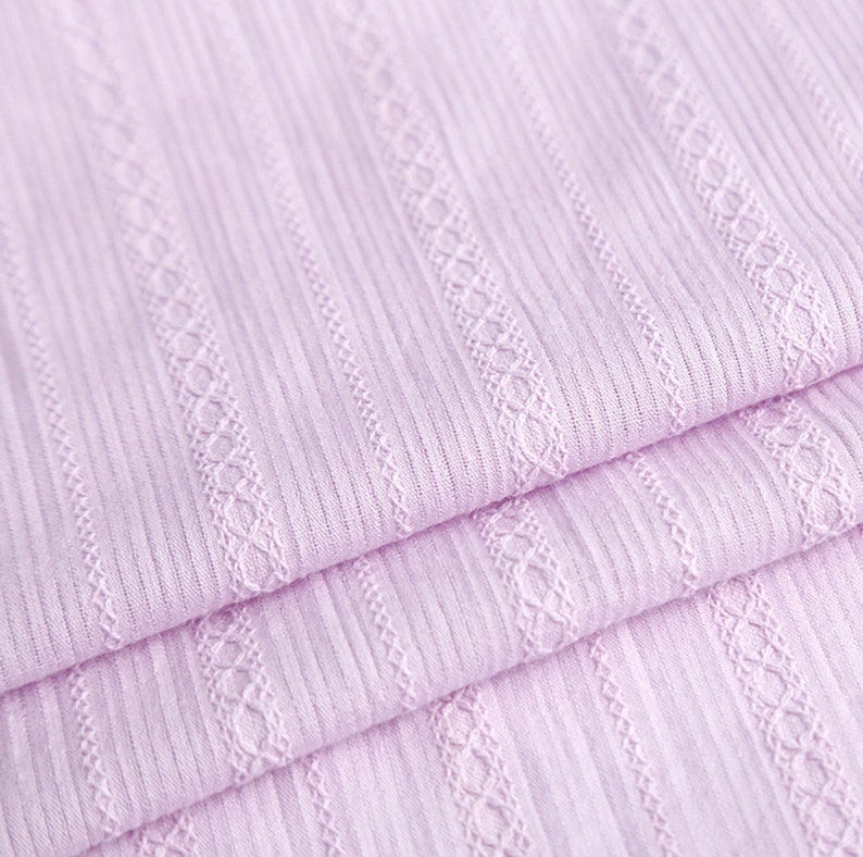 May include: Close-up of a soft, pale pink fabric with a ribbed texture and a delicate, woven pattern. The fabric is folded in layers, showcasing the intricate details of the design.