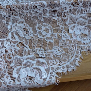 May include: White lace fabric with a floral pattern. The lace has a scalloped edge and a delicate, frayed finish.