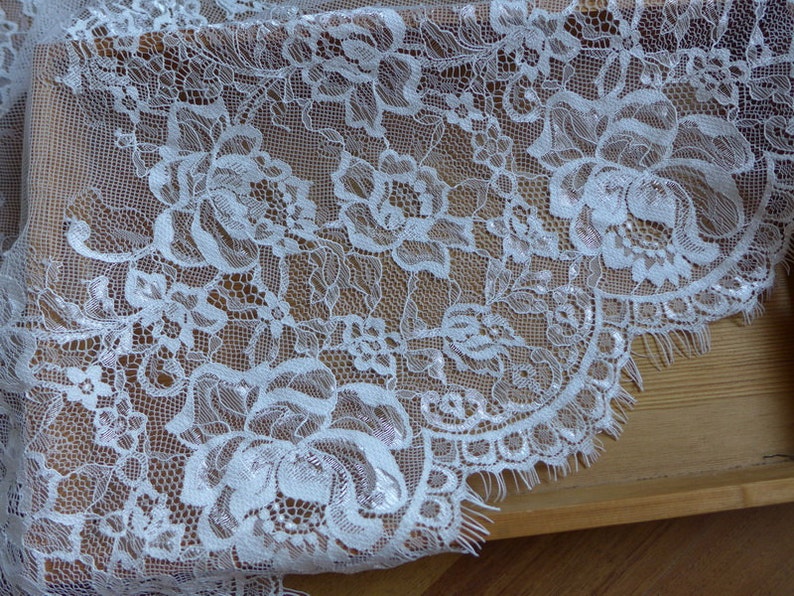 May include: White lace fabric with a floral pattern. The lace has a scalloped edge and a delicate, frayed finish.