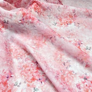 May include: A sheer, lightweight fabric featuring a delicate, crinkled texture and an all-over floral print. The pattern consists of soft pink and coral-coloured blossoms with subtle hints of purple and grey foliage scattered across a white background.