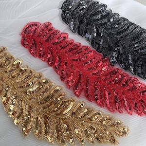 May include: Three sequined feather appliques in gold, red, and black are displayed. The feathers are intricately detailed with small, shiny sequins. They are arranged on a white fabric background. Ideal for embellishing garments or craft projects.