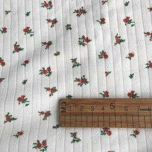 May include: Close-up of white fabric with a ribbed texture and a pattern of small red roses and green leaves. A wooden ruler is placed on the fabric, showing measurements in centimeters.