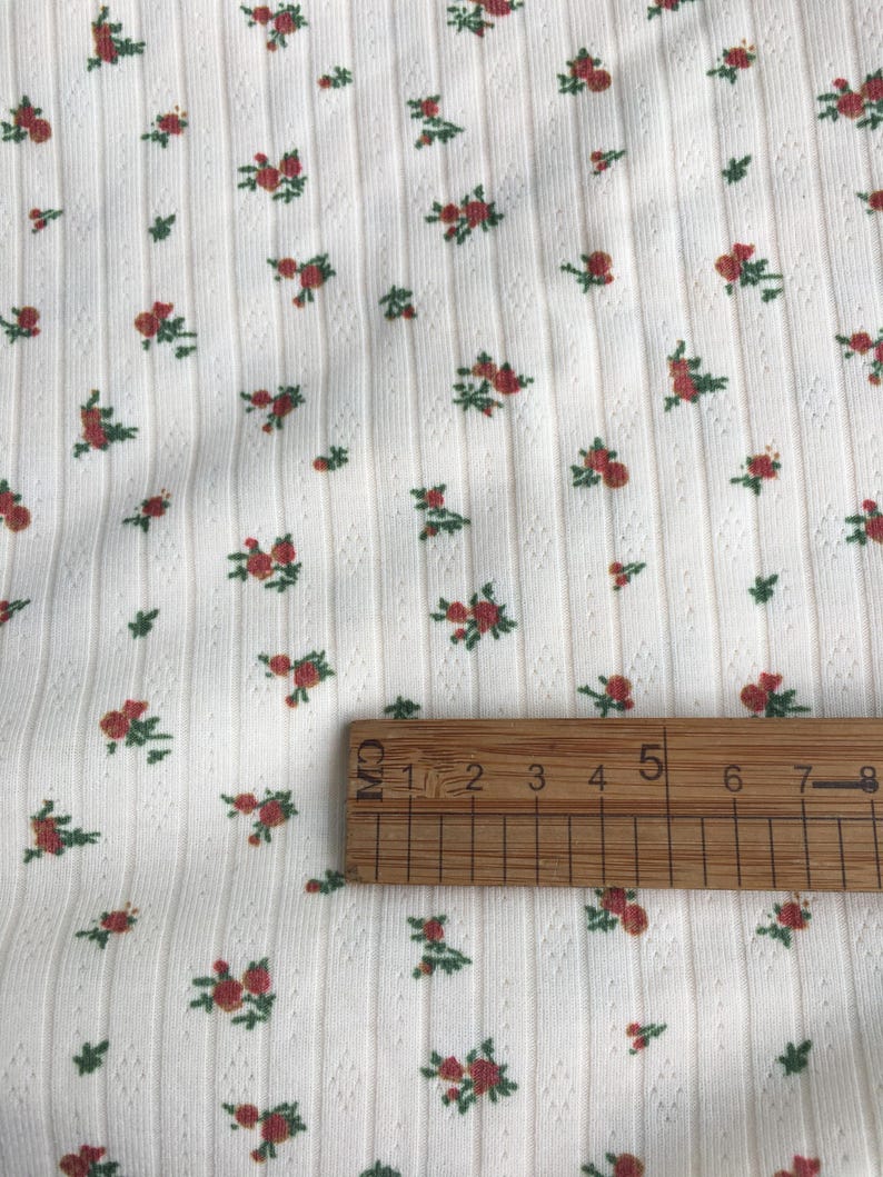 May include: Close-up of white fabric with a ribbed texture and a pattern of small red roses and green leaves. A wooden ruler is placed on the fabric, showing measurements in centimeters.
